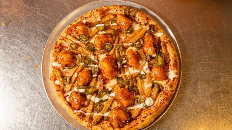 Pizza topped with crispy chicken, french fries, sliced jalapeños, and white sauce on a metal tray.