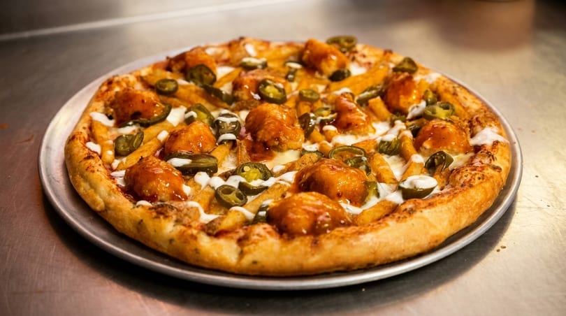 A pizza topped with crispy fries, boneless chicken wings, sliced jalapeños, and white sauce.