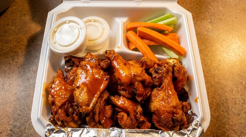 Takeout container filled with saucy chicken wings, celery, carrots, and two dipping sauces.