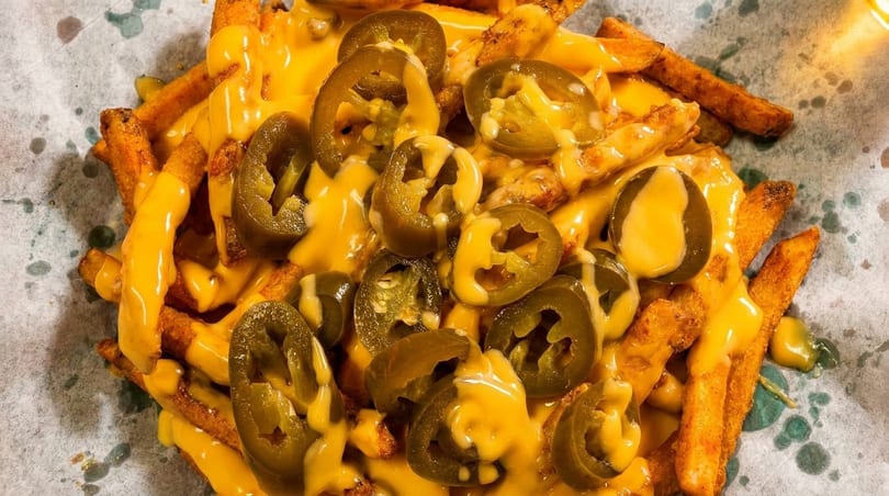 Golden french fries generously covered in melted cheese and sliced jalapeños on a paper-lined tray.