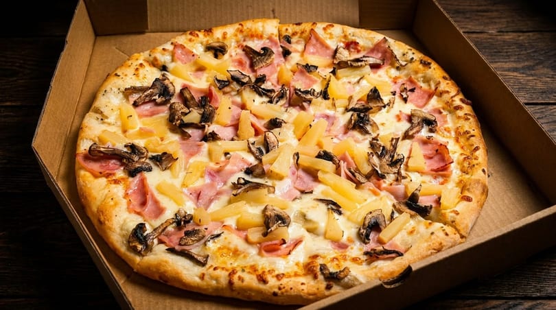 A whole pizza with ham, pineapple, and mushrooms in a cardboard delivery box.
